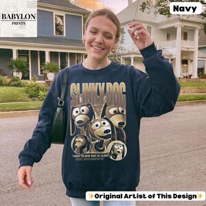 May include: Navy blue sweatshirt featuring the text "SLINKY DOG" in gold lettering, with a graphic of the Slinky Dog character from the Toy Story movie. The sweatshirt also includes the text "I KNEW YOU WERE RIGHT ALL ALONG WOODY NEVER DOUBTED YA".