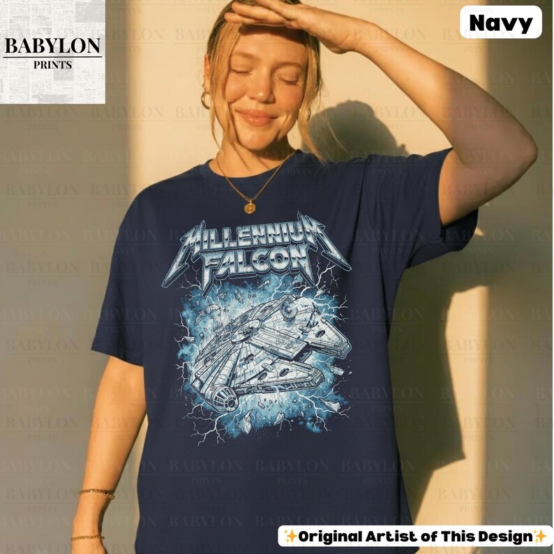 May include: Navy blue t-shirt featuring a graphic of the Millennium Falcon spaceship with the text "Millennium Falcon" in a metallic font. The design includes lightning bolts and is set against a blue background. The shirt is from Babylon Prints.