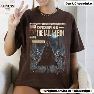 May include: Dark chocolate brown t-shirt featuring a comic book-style graphic from Star Wars. The design includes the text "ORDER 66 - THE FALL OF THE JEDI" and an illustration of Darth Vader and Stormtroopers. The shirt has a crew neck.