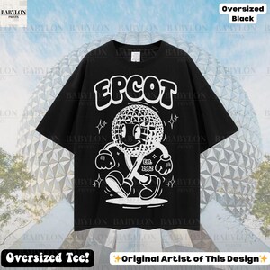 Pu&ograve; includere: T-shirt nero oversize con una grafica bianca di un personaggio geometrico sorridente e la scritta "EPCOT". La grafica include il testo "Est. 1982". La maglietta ha anche il testo "Oversized Tee!" e "Original Artist of This Design".
