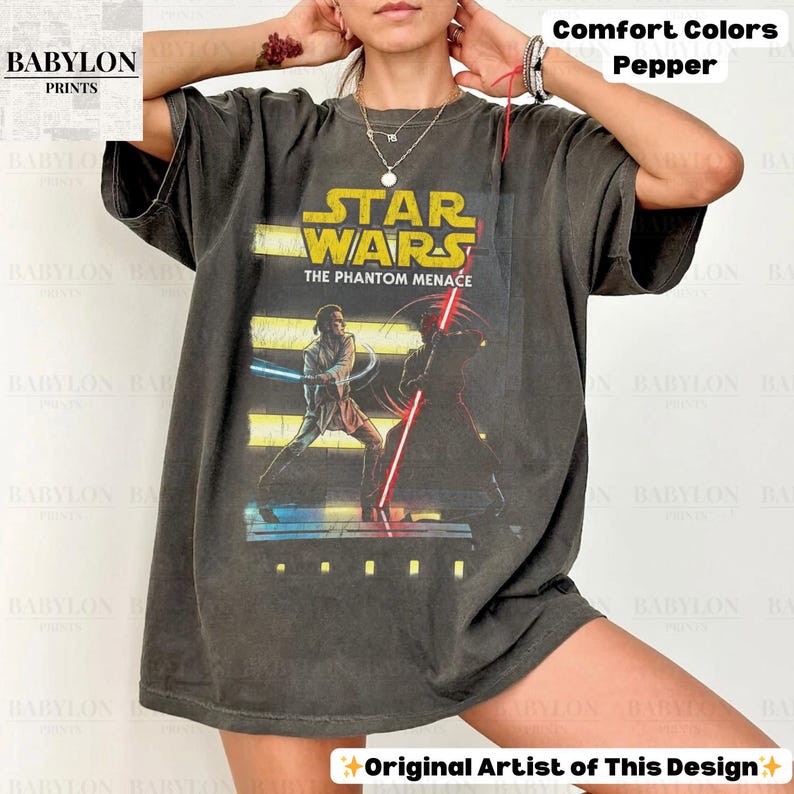Puede incluir: Una camiseta gris oscuro con un gr&aacute;fico de Star Wars. El dise&ntilde;o incluye el texto "STAR WARS THE PHANTOM MENACE" e im&aacute;genes de personajes con sables de luz. La camiseta est&aacute; etiquetada como "Comfort Colors Pepper" y "Original Artist of This Design."