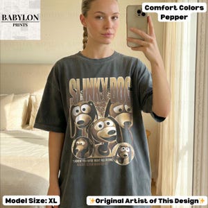 May include: A dark gray t-shirt featuring the text "SLINKY DOG" above a graphic of the Slinky Dog character from Toy Story. The shirt also includes the text "I KNEW YOU WERE RIGHT ALL ALONG WOODY NEVER DOUBTED YOU". The shirt is a comfort color in pepper.