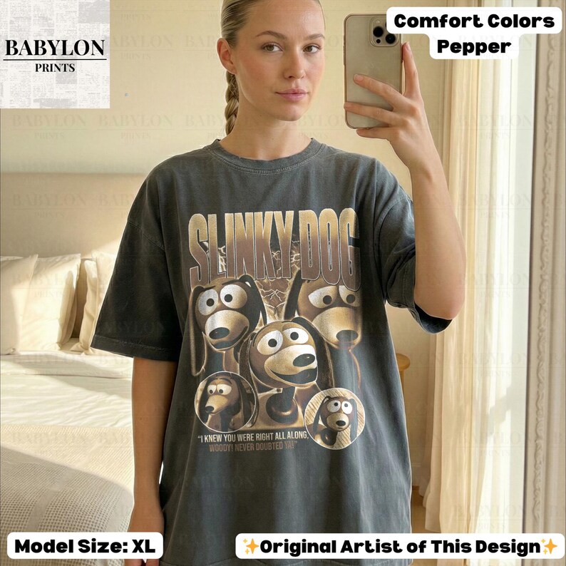 May include: A dark gray t-shirt featuring the text "SLINKY DOG" above a graphic of the Slinky Dog character from Toy Story. The shirt also includes the text "I KNEW YOU WERE RIGHT ALL ALONG WOODY NEVER DOUBTED YOU". The shirt is a comfort color in pepper.