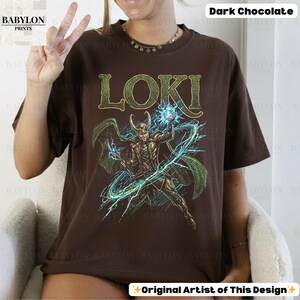 May include: Dark chocolate-colored t-shirt featuring the word "LOKI" in green, above a detailed illustration of a character with green cape and horns, wielding blue energy. The shirt is from Babylon Prints.