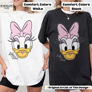 May include: White and black t-shirts featuring a Daisy Duck graphic. The design includes a pink bow, eyelashes, and the text "Daisy Duck". The shirts are labeled "Comfort Colors" and "Original Artist of This Design".
