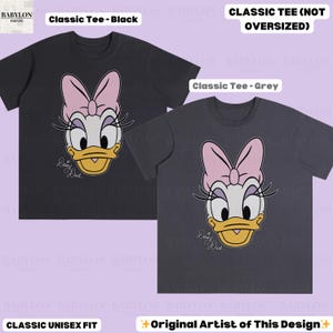 Puede incluir: Dos camisetas cl&aacute;sicas unisex, una negra y otra gris, cada una con un dise&ntilde;o de Daisy Duck. El dise&ntilde;o incluye un lazo rosa, un pico amarillo y el texto "Daisy Duck". Las camisetas est&aacute;n etiquetadas como "Classic Tee" y "Original Artist of This Design".