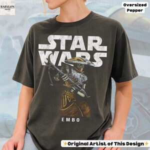 Puede incluir: Camiseta gris oscuro de gran tama&ntilde;o con un gr&aacute;fico de Star Wars y el texto "STAR WARS" en blanco. La camiseta muestra una ilustraci&oacute;n del personaje Embo, sosteniendo un arma. El texto "EMBO" est&aacute; debajo. La camiseta est&aacute; etiquetada como "Oversized Pepper" y "Original Artist of This Design."