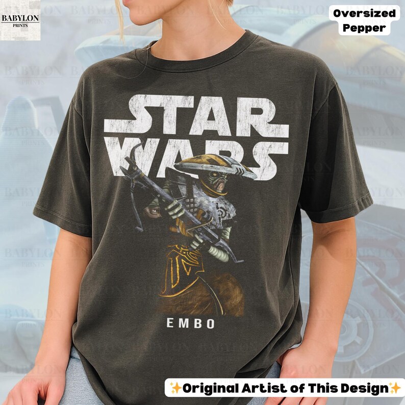 Puede incluir: Camiseta gris oscuro de gran tama&ntilde;o con un gr&aacute;fico de Star Wars y el texto "STAR WARS" en blanco. La camiseta muestra una ilustraci&oacute;n del personaje Embo, sosteniendo un arma. El texto "EMBO" est&aacute; debajo. La camiseta est&aacute; etiquetada como "Oversized Pepper" y "Original Artist of This Design."