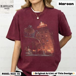 May include: Maroon t-shirt with a graphic of a building and fountain, and the text "GUSTEAU'S". The shirt also has the text "BABYLON PRINTS" and "Original Artist of This Design". The model is wearing a size XL.