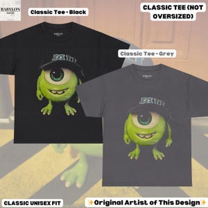 K&ouml;nnte beinhalten: Zwei klassische Unisex-T-Shirts, eines in Schwarz und eines in Grau, jeweils mit einer Cartoonfigur mit einem gro&szlig;en gr&uuml;nen Auge, die einen Hut tr&auml;gt. Das schwarze Shirt hat den Text "Classic Tee - Black" und das graue Shirt den Text "Classic Tee - Grey". Der Text "Original Artist of This Design" ist ebenfalls vorhanden.