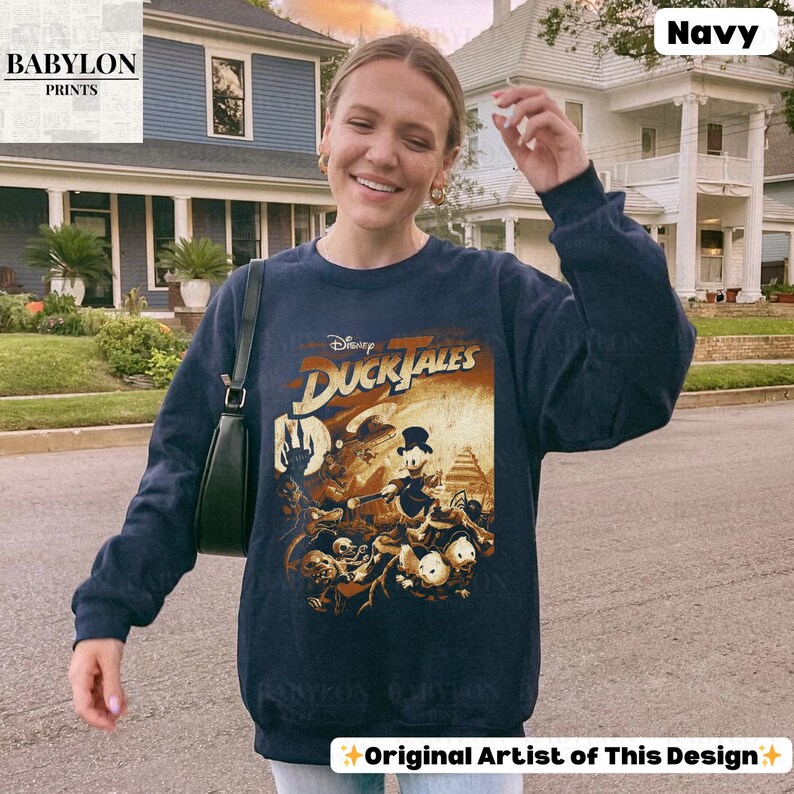 May include: Navy blue sweatshirt featuring a Disney DuckTales graphic. The graphic is in shades of brown, orange, and white, with the text "DuckTales" in a stylized font. The sweatshirt has a crew neck and long sleeves. The words "Original Artist of This Design" are at the bottom.