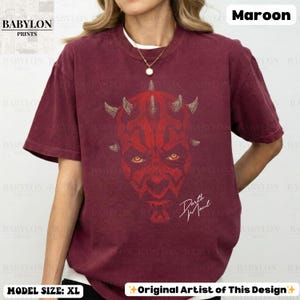 Może przedstawiać: Bordowa koszulka z czerwoną grafiką twarzy Darth Maula, z rogami i ż&oacute;łtymi oczami. Tekst "Darth Maul" jest napisany białą czcionką. Koszulka ma tekst "BABYLON PRINTS" w lewym g&oacute;rnym rogu.