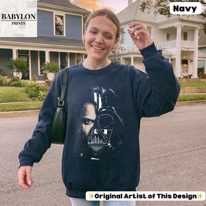 May include: Navy blue sweatshirt featuring a graphic of Darth Vader's face. The design is split, showing both the character's face and helmet. The text "Original Artist of This Design" is at the bottom. The sweatshirt is worn by a person.