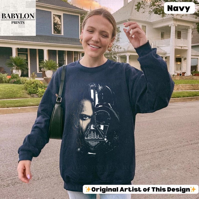 May include: Navy blue sweatshirt featuring a graphic of Darth Vader's face. The design is split, showing both the character's face and helmet. The text "Original Artist of This Design" is at the bottom. The sweatshirt is worn by a person.