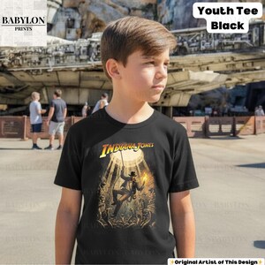 May include: Black youth tee featuring an Indiana Jones graphic. The design shows Indiana Jones in a well, holding a torch and rope. The shirt has the text "Youth Tee Black" in the upper right corner. The Babylon Prints logo is in the upper left corner.
