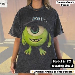 K&ouml;nnte beinhalten: Ein dunkelgraues T-Shirt mit einer gro&szlig;en Grafik von Mike Wazowski aus dem Film Die Monster AG. Das Design enth&auml;lt das Wort "MIU" in Blau und Wei&szlig;. Das Shirt ist eine Premium-Waschung in Pfefferfarbe.