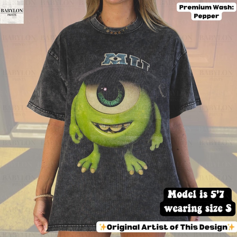 K&ouml;nnte beinhalten: Ein dunkelgraues T-Shirt mit einer gro&szlig;en Grafik von Mike Wazowski aus dem Film Die Monster AG. Das Design enth&auml;lt das Wort "MIU" in Blau und Wei&szlig;. Das Shirt ist eine Premium-Waschung in Pfefferfarbe.