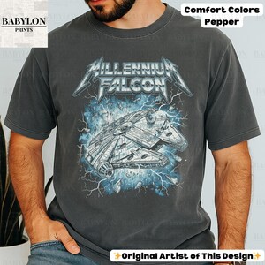 May include: A dark grey t-shirt featuring a graphic of the Millennium Falcon spaceship with the text "MILLENNIUM FALCON" in a stylized font. The design includes blue lightning and the words "Comfort Colors Pepper". The shirt is from "BABYLON PRINTS".