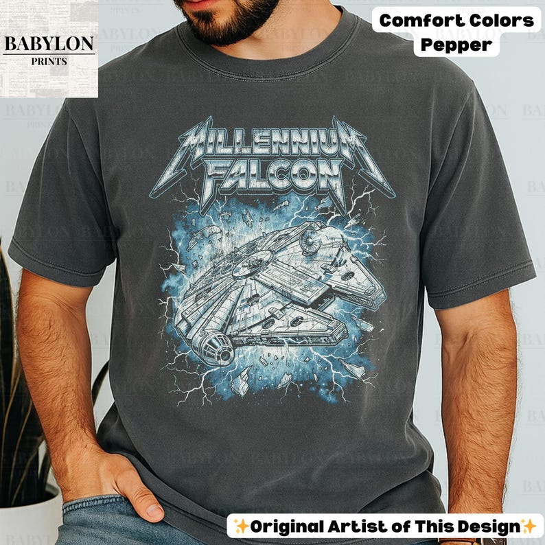 May include: A dark grey t-shirt featuring a graphic of the Millennium Falcon spaceship with the text "MILLENNIUM FALCON" in a stylized font. The design includes blue lightning and the words "Comfort Colors Pepper". The shirt is from "BABYLON PRINTS".