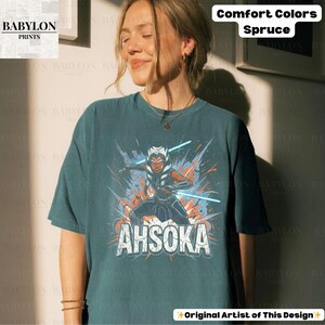 Może przedstawiać: Tealowa koszulka z grafiką Ahsoki Tano z mieczami świetlnymi i napisem "AHSOKA". Koszulka zawiera r&oacute;wnież tekst "Comfort Colors Spruce" i "Original Artist of This Design".