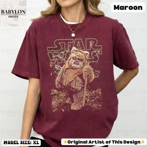 May include: Maroon t-shirt featuring a Star Wars graphic. The design includes the Star Wars logo and an Ewok character holding a staff. The shirt has short sleeves and a crew neck. The text "BABYLON PRINTS" is visible.
