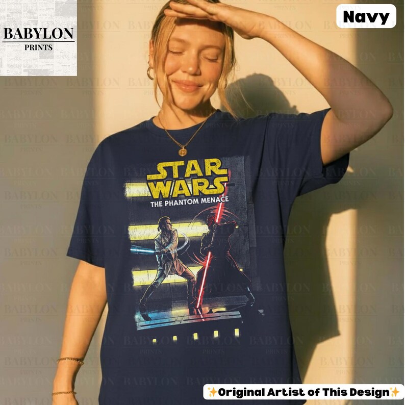 Puede incluir: Camiseta azul marino con un dise&ntilde;o de Star Wars: La Amenaza Fantasma. El dise&ntilde;o incluye el logotipo de Star Wars y personajes con sables de luz. Tambi&eacute;n se ve el texto "Original Artist of This Design".