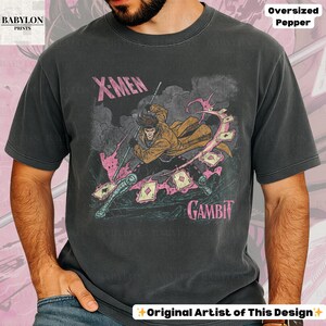 May include: Dark grey oversized t-shirt featuring a graphic of the X-Men character Gambit. The design includes Gambit wielding a staff and playing cards, with the text "X-Men" and "Gambit" in pink. The shirt is labeled "Oversized Pepper" and "Original Artist of This Design."