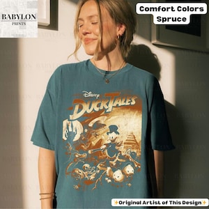 May include: A teal-colored t-shirt featuring a graphic from Disney's DuckTales. The design includes characters like Scrooge McDuck and Huey, Dewey, and Louie. The shirt also has the text "Comfort Colors Spruce" and "Original Artist of This Design".
