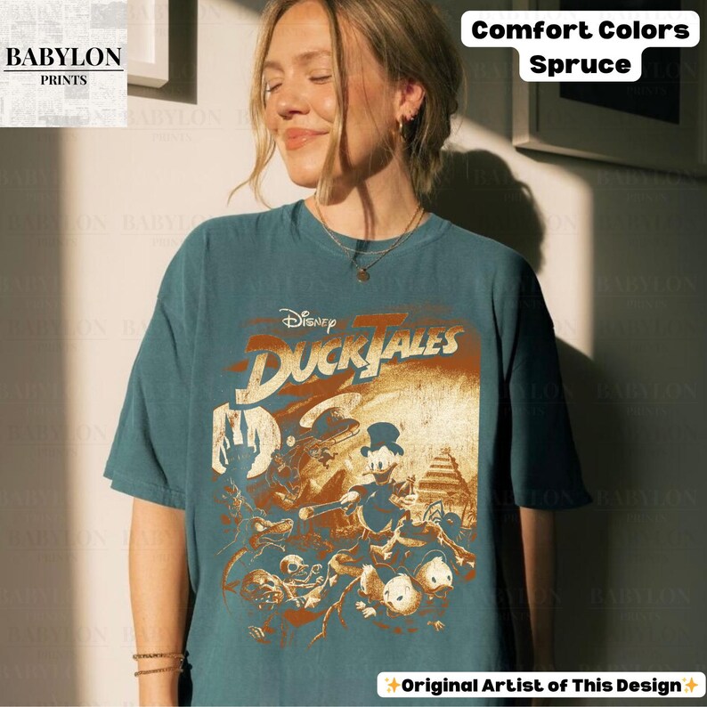 May include: A teal-colored t-shirt featuring a graphic from Disney's DuckTales. The design includes characters like Scrooge McDuck and Huey, Dewey, and Louie. The shirt also has the text "Comfort Colors Spruce" and "Original Artist of This Design".