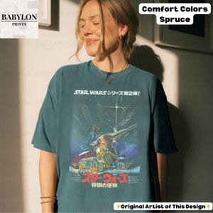 May include: A spruce-colored t-shirt featuring a Star Wars movie poster design with Japanese text. The graphic includes images of characters and a spaceship. The shirt has the text "STAR WARS シリーズ第2弾!" and "スターウォーズ 帝国の逆襲".