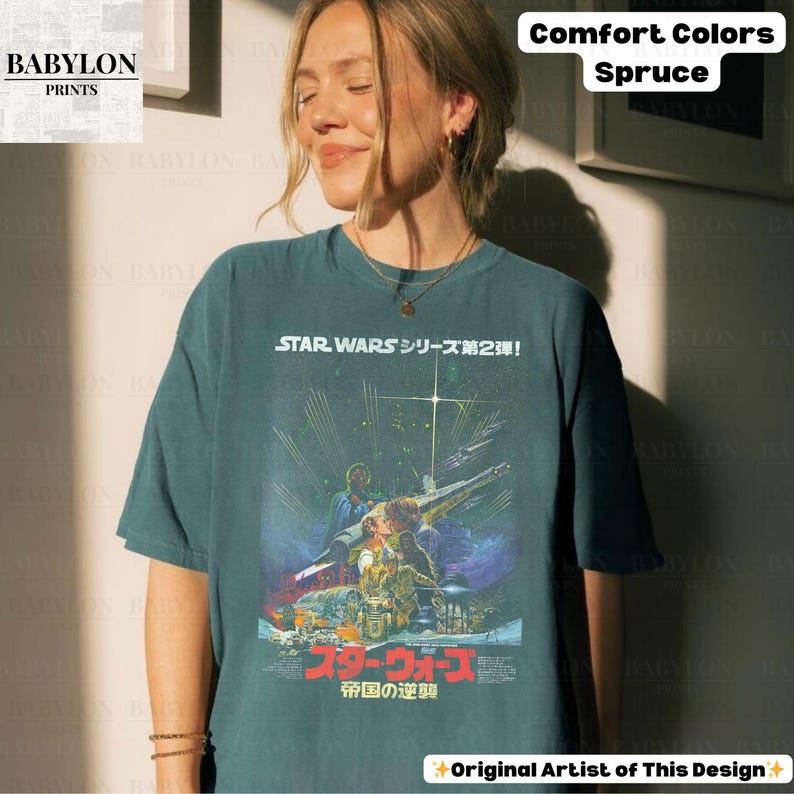 May include: A spruce-colored t-shirt featuring a Star Wars movie poster design with Japanese text. The graphic includes images of characters and a spaceship. The shirt has the text "STAR WARS シリーズ第2弾!" and "スターウォーズ 帝国の逆襲".