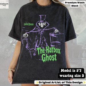 Puede incluir: Una camiseta negra con efecto lavado al &aacute;cido que presenta un gr&aacute;fico de The Hatbox Ghost de The Haunted Mansion. El personaje lleva un sombrero de copa y sostiene un bast&oacute;n y una sombrerera. El texto dice The Haunted Mansion y The Hatbox Ghost en verde.