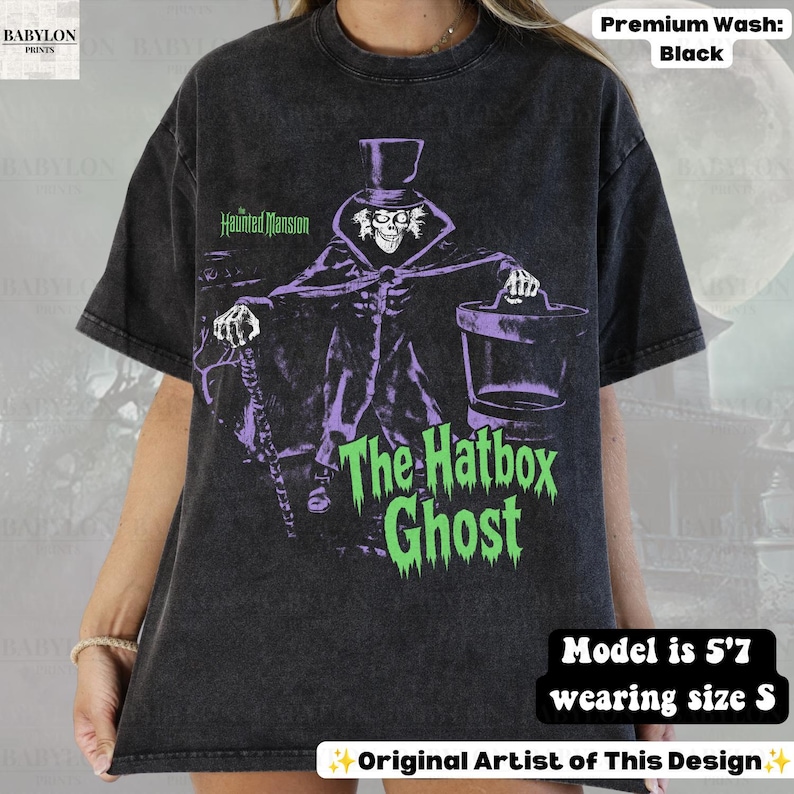 Puede incluir: Una camiseta negra con efecto lavado al &aacute;cido que presenta un gr&aacute;fico de The Hatbox Ghost de The Haunted Mansion. El personaje lleva un sombrero de copa y sostiene un bast&oacute;n y una sombrerera. El texto dice The Haunted Mansion y The Hatbox Ghost en verde.