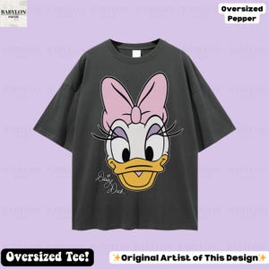 Puede incluir: Camiseta gris oscuro de gran tama&ntilde;o con un gran gr&aacute;fico de Daisy Duck con un lazo rosa, pesta&ntilde;as y un pico amarillo. El texto "Daisy Duck" est&aacute; escrito en cursiva. La camiseta tiene el texto "Oversized Tee!" y "Original Artist of This Design".