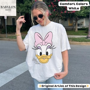 May include: White Comfort Colors t-shirt featuring a Daisy Duck graphic with a pink bow, eyelashes, and a yellow beak. The shirt includes the text "Daisy Duck" and "Original Artist of This Design."