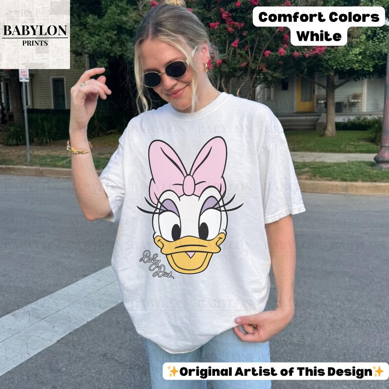 May include: White Comfort Colors t-shirt featuring a Daisy Duck graphic with a pink bow, eyelashes, and a yellow beak. The shirt includes the text "Daisy Duck" and "Original Artist of This Design."