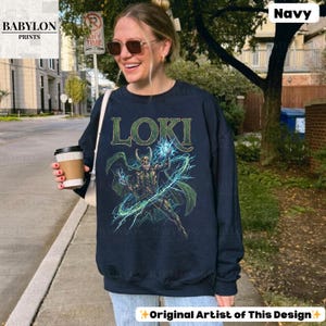 May include: Navy blue sweatshirt featuring a graphic of Loki with the word "LOKI" above. The design includes green and blue accents. The sweatshirt is worn by a person outdoors.