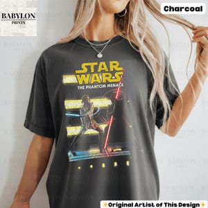Puede incluir: Camiseta de color carb&oacute;n con un gr&aacute;fico de Star Wars. El dise&ntilde;o incluye el texto "STAR WARS THE PHANTOM MENACE" en amarillo y rojo, con una escena de dos personajes con sables de luz. El logotipo de Babylon Prints es visible.
