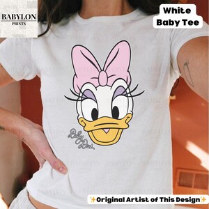 May include: White baby tee featuring a cartoon illustration of Daisy Duck with a pink bow, eyelashes, and a yellow beak. The text "Daisy Duck" is below the illustration. The shirt has the text "White Baby Tee" and "Original Artist of This Design".