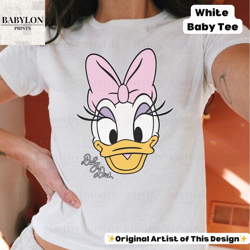 May include: White baby tee featuring a cartoon illustration of Daisy Duck with a pink bow, eyelashes, and a yellow beak. The text "Daisy Duck" is below the illustration. The shirt has the text "White Baby Tee" and "Original Artist of This Design".
