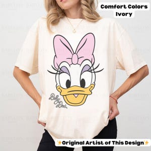 May include: Ivory Comfort Colors t-shirt featuring a large graphic of Daisy Duck's face. The design includes a pink bow, eyelashes, and the words "Daisy Duck" in script. The shirt is a short-sleeved, casual style.