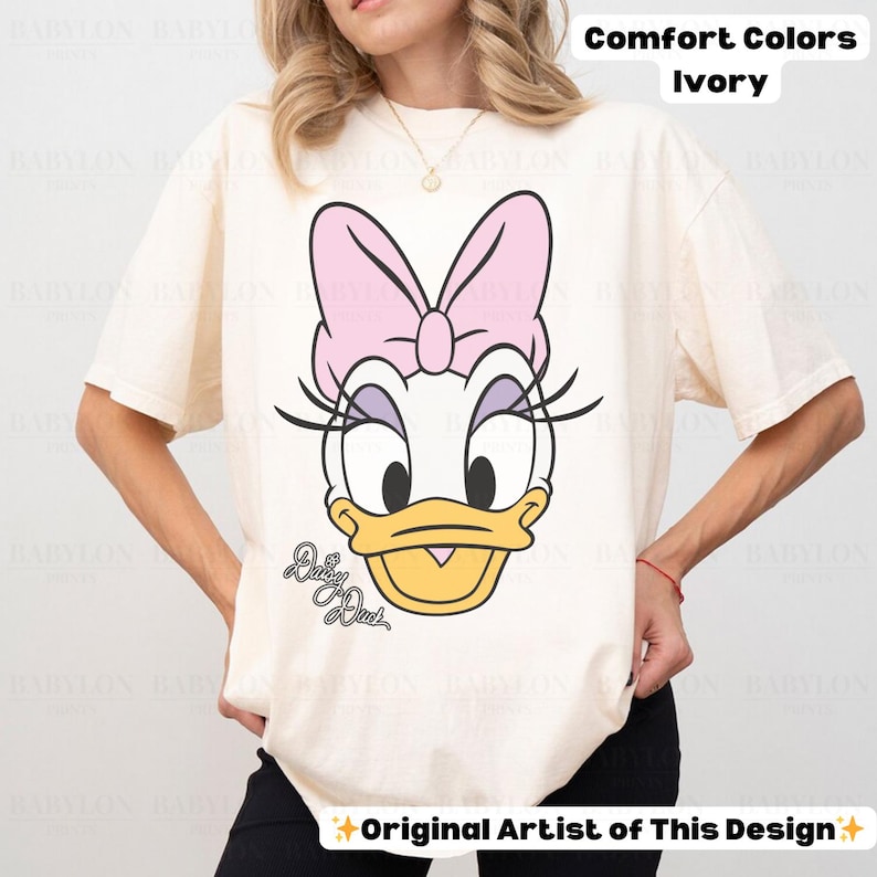 May include: Ivory Comfort Colors t-shirt featuring a large graphic of Daisy Duck's face. The design includes a pink bow, eyelashes, and the words "Daisy Duck" in script. The shirt is a short-sleeved, casual style.