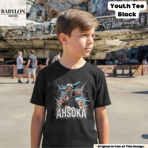 Może przedstawiać: Czarny t-shirt dla młodzieży z grafiką Ahsoki Tano z mieczami świetlnymi i napisem "AHSOKA" w kolorze białym. Koszulka ma kr&oacute;tkie rękawy i okrągły dekolt. Słowa "Youth Tee Black" są wydrukowane na g&oacute;rze po prawej stronie.