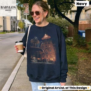 May include: Navy blue sweatshirt with a graphic of a building and fountain, and the text "GUSTEAU'S". The long-sleeved, crew neck sweatshirt also features the text "Original Artist of This Design".