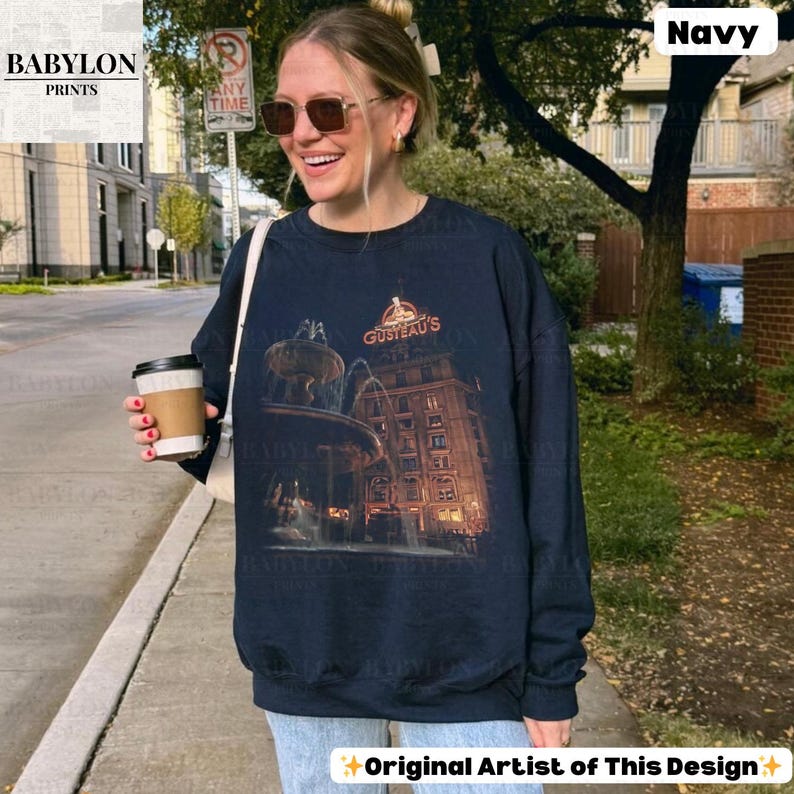 May include: Navy blue sweatshirt with a graphic of a building and fountain, and the text "GUSTEAU'S". The long-sleeved, crew neck sweatshirt also features the text "Original Artist of This Design".