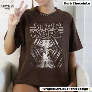 May include: Dark chocolate brown Star Wars t-shirt with a white graphic. The graphic features the Star Wars logo and a detailed image of a hallway with a figure. The shirt is a relaxed fit.