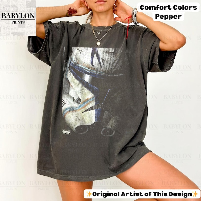 May include: A charcoal gray short-sleeve t-shirt featuring a large graphic print of a white and blue clone trooper helmet. The design includes the text "CLONE WARS" in small white letters below the helmet. The shirt has a loose, relaxed fit.