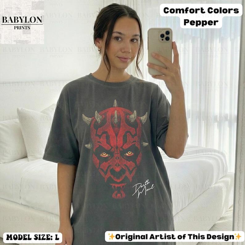 Może przedstawiać: Ciemnoszara koszulka z czerwono-czarną grafiką Darth Maula. Koszulka ma napis "Darth Maul" w białym skrypcie. Oznaczona jako "Comfort Colors Pepper" i "Original Artist of This Design". Modelka nosi rozmiar L.
