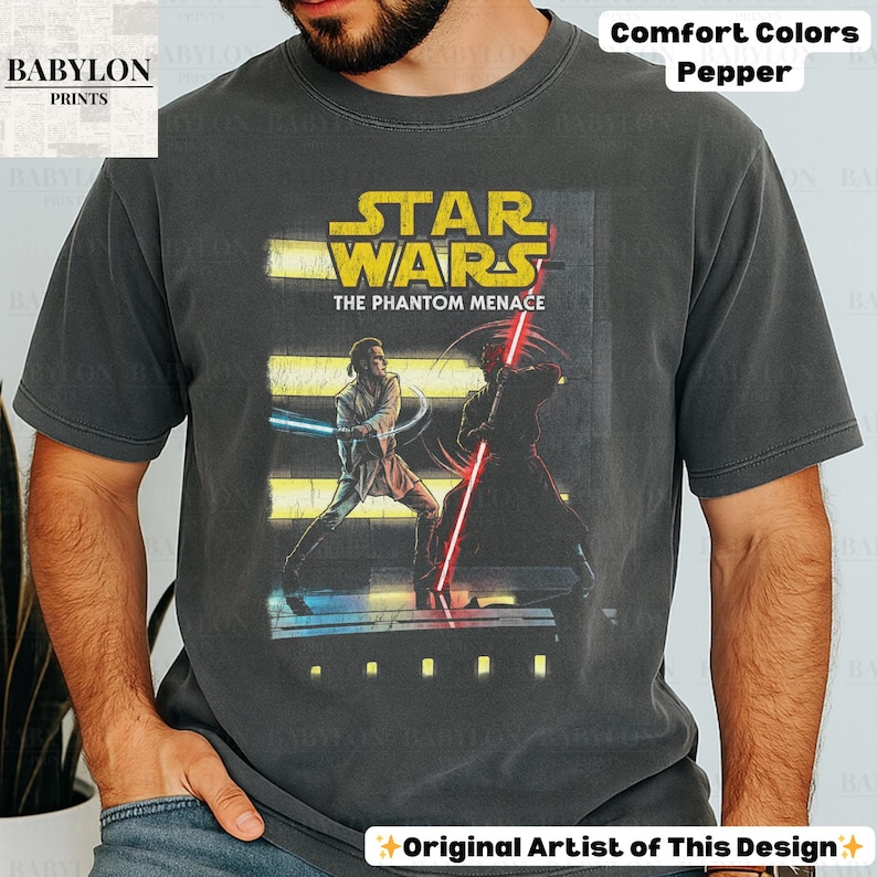 Puede incluir: Camiseta gris oscuro con un gr&aacute;fico de Star Wars. El dise&ntilde;o incluye el texto "STAR WARS THE PHANTOM MENACE" en amarillo, y una escena con dos personajes con sables de luz. La camiseta es "Comfort Colors Pepper".