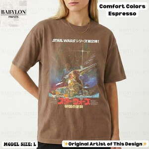 May include: Espresso-colored t-shirt featuring a Star Wars movie poster design. The graphic includes Japanese text and a vintage aesthetic. The shirt is labeled as Comfort Colors and the model is wearing a size L.